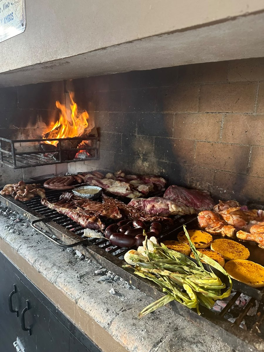 An asado on a brick grill: steaks, sausages, blood sausage, grilled vegetables and polenta, with a fire burning behind